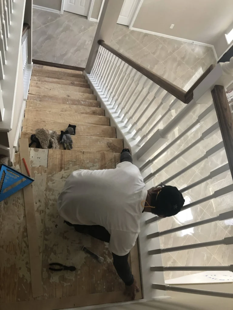 Stair remodel in progress in Cypress TX with carpet removed, exposed plywood sub-treads, and a carpenter fitting materials along the existing white baluster railing.