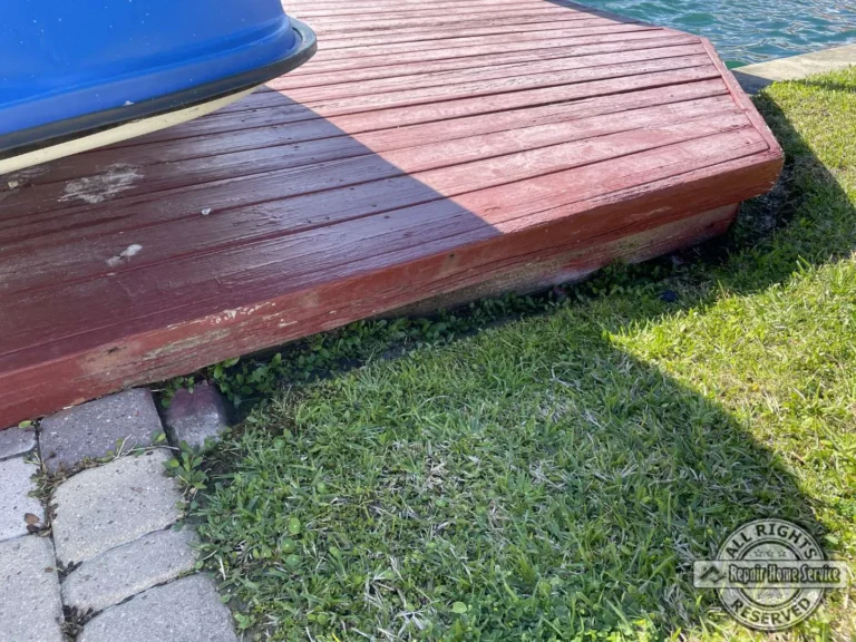 Weathered waterfront deck boards in Houston TX 77065 with chipped red stain, stone pavers, grass, and a small boat on the dock before refinishing.