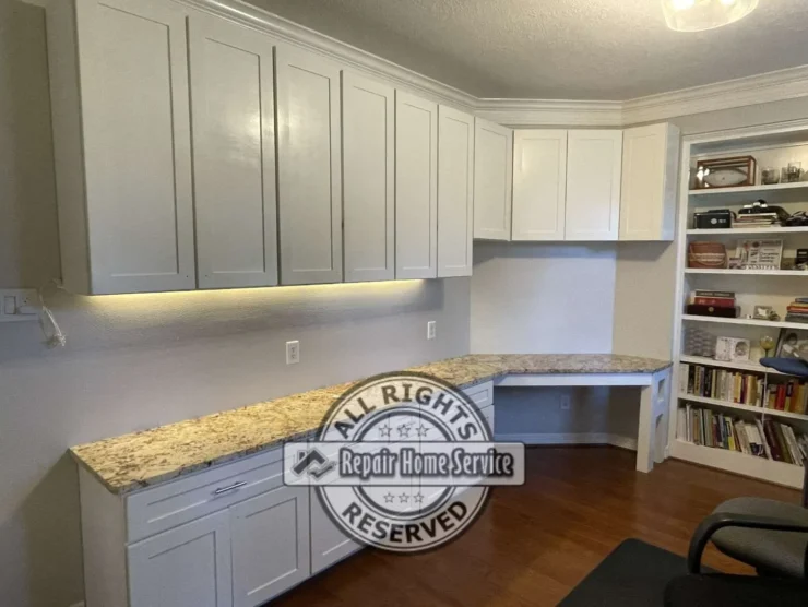 Completed custom craft room built-ins in Cypress TX with white shaker cabinets, granite countertop, under-cabinet lighting, corner desk, and built-in bookcase filled with books and supplies.