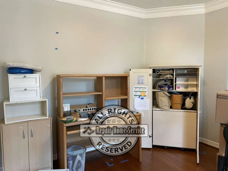 Before photo of cluttered Cypress craft room with mismatched oak desk, small wood cabinet, and open storage unit marked with blue tape for new built-in cabinet layout.