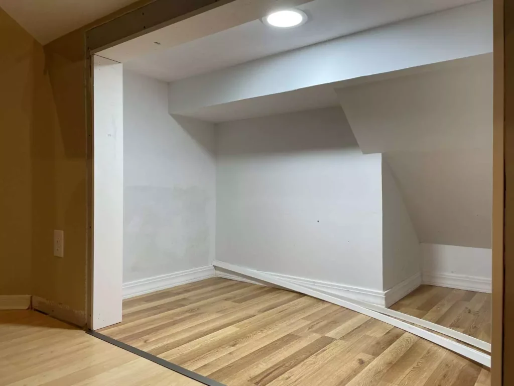 Finished white-painted under-stairs alcove in a Houston TX home with light laminate flooring, recessed ceiling light, and loose trim pieces before custom hidden storage installation.