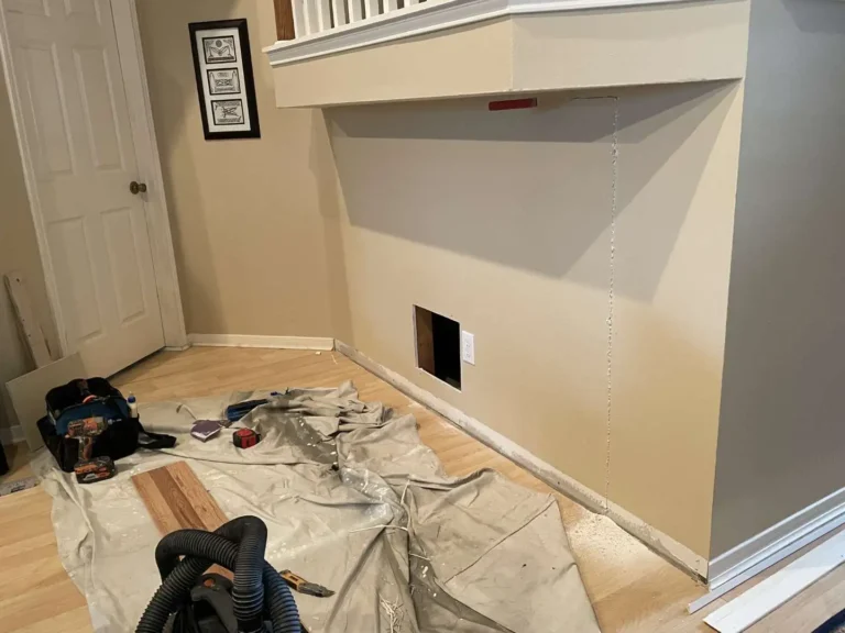 Vertical drywall cut marked under a staircase overhang in Houston TX during custom hidden storage layout with drop cloth, tools, and laminate flooring protected on site.