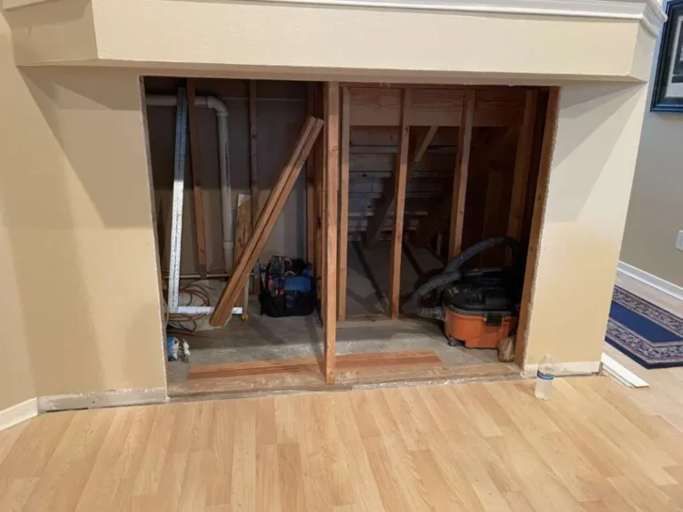Open wall cavity under stairs in Houston TX showing exposed studs, plumbing pipe, shop vacuum, and carpentry tools during hidden storage rough-in work.