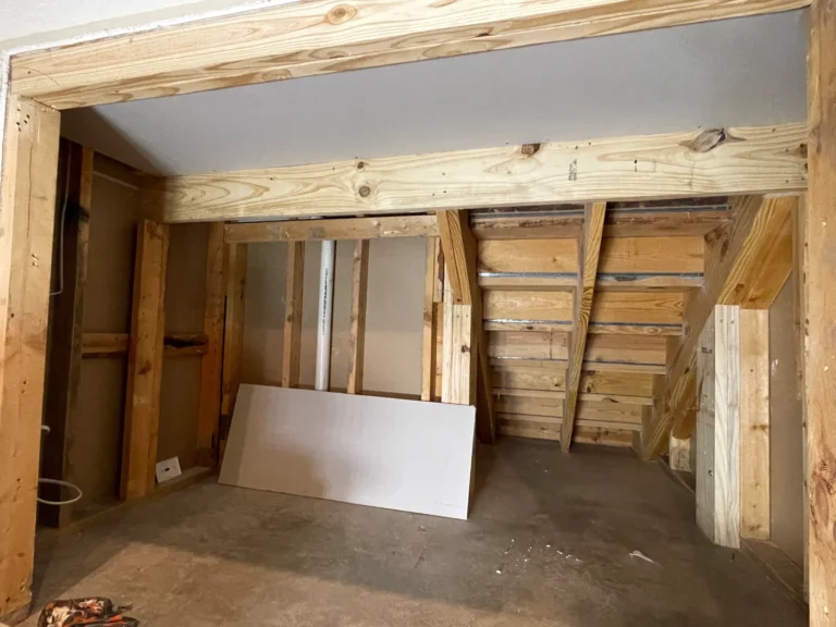 Raw wood framing inside an under-stair opening in Houston TX with concrete floor, vertical studs, and staircase stringers visible before hidden storage build-out.
