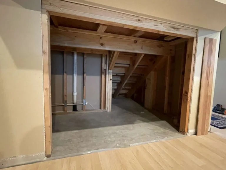 Framed rectangular opening under a staircase in a Houston TX living area with exposed interior studs, laminate flooring, and unfinished concrete inside the cavity.