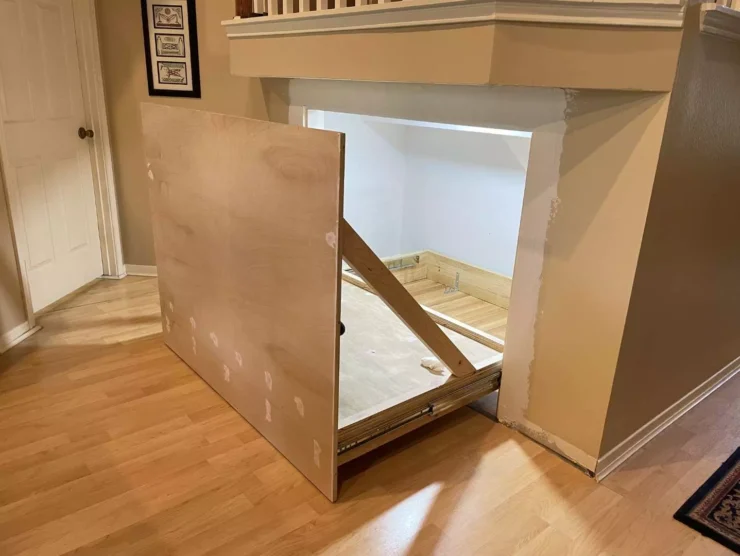 Sliding hidden storage unit partially pulled out under stairs in Houston TX showing plywood face panel, drawer slides, white-painted interior, and staircase trim above.