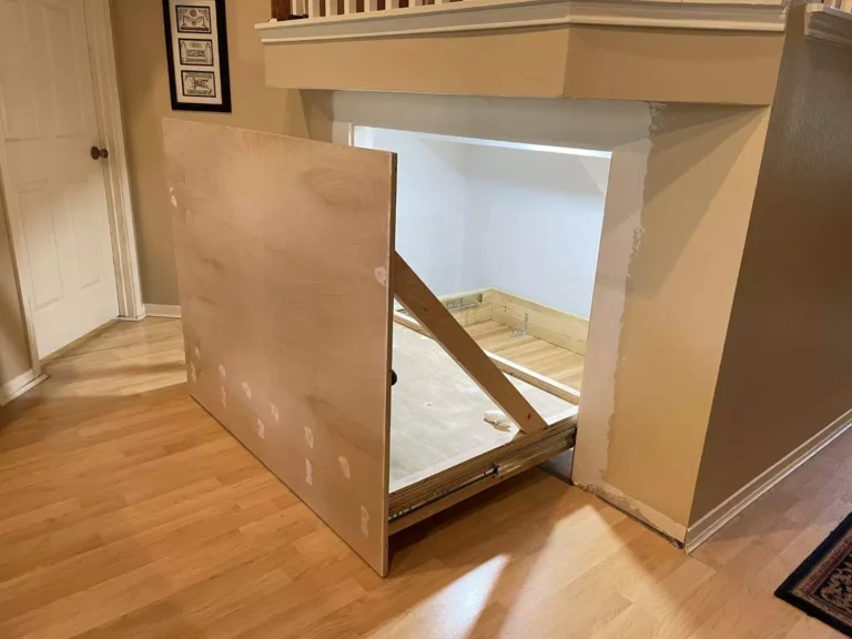 Sliding hidden storage unit partially pulled out under stairs in Houston TX showing plywood face panel, drawer slides, white-painted interior, and staircase trim above.