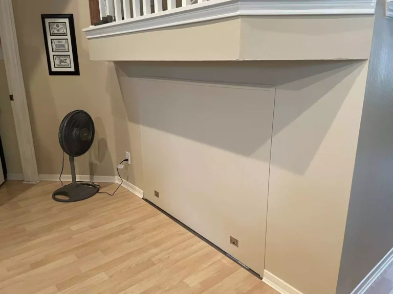 Closed hidden storage door under staircase in Houston TX blending flush with beige wall paint and stainless recessed finger pulls at the bottom edge.