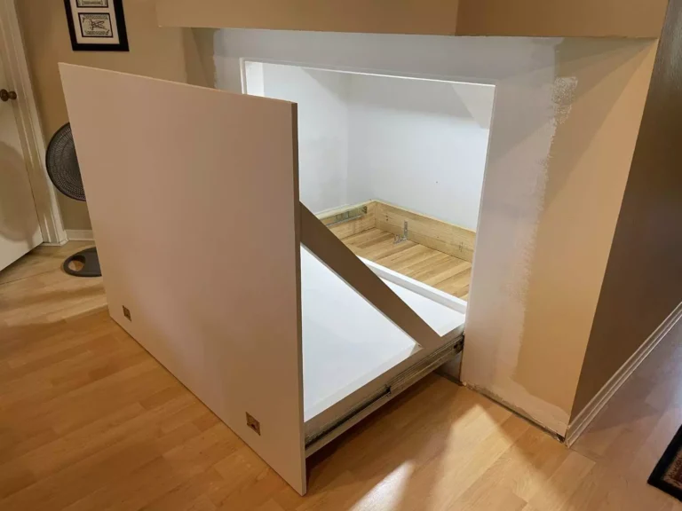 Custom hidden storage panel pulled open under stairs in Houston TX revealing primed door face, metal slides, lit white interior, and laminate floor inside the compartment.