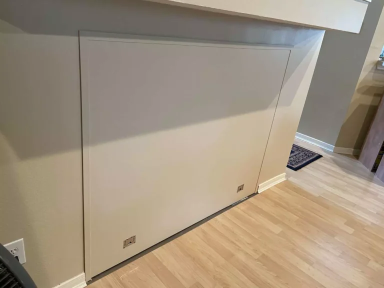 Close-up of seamless hidden storage door in Houston TX with flat painted finish, tight trim joints, and two stainless recessed finger pulls above laminate flooring.
