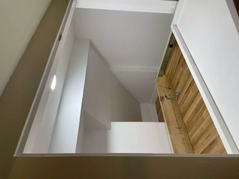 Interior view of open hidden under-stair storage in Houston TX with blond plywood pull-out platform, laminate surface, LED puck light, metal slides, and white flat-painted walls.