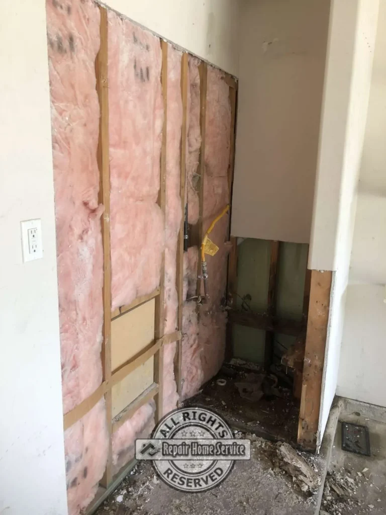 Utility closet with damaged drywall removed, exposed wall studs, pink insulation, plumbing lines, and debris from water heater leak repair.