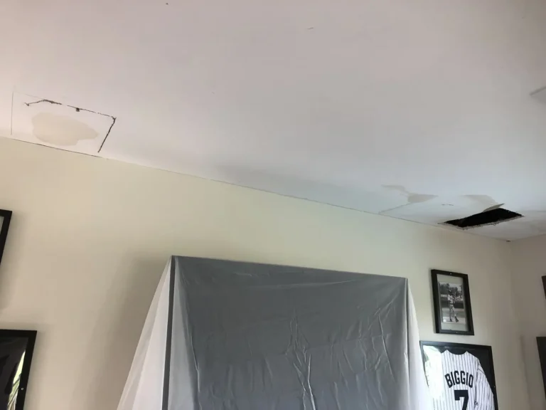 Living room ceiling before repair with water-damaged drywall patches, open ceiling hole, wall pictures, covered furniture, and HVAC vent area.