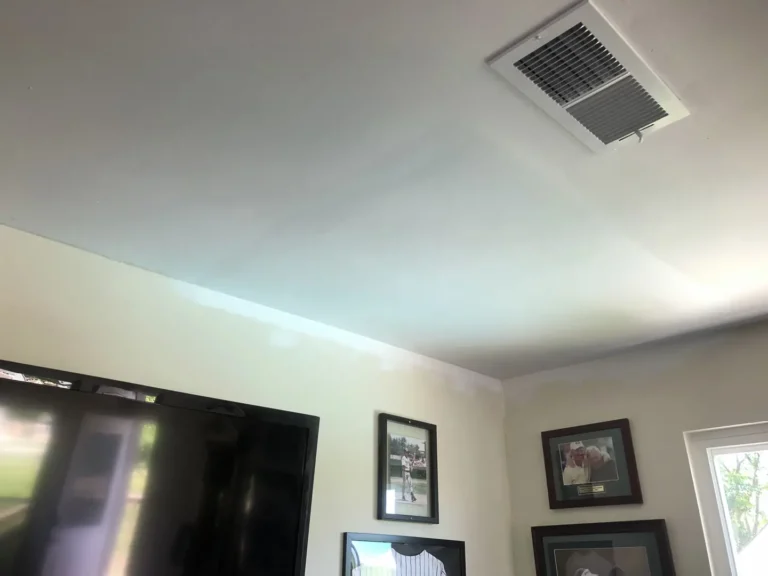 Living room ceiling after Sheetrock repair with smooth patched finish, HVAC vent, framed wall art, TV, and repaired ceiling surface ready for paint.