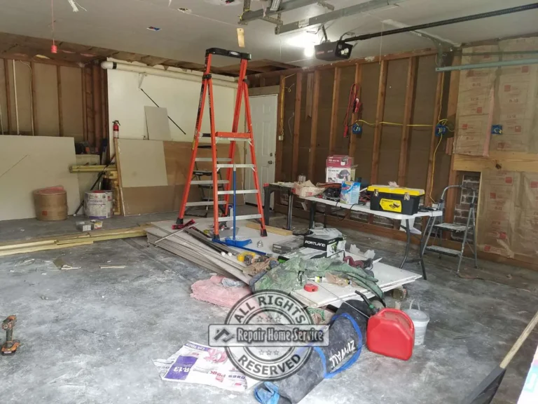 Garage remodel in progress with exposed wall studs, insulation, ladder, tools, drywall sheets, garage door opener, and ceiling repair work.