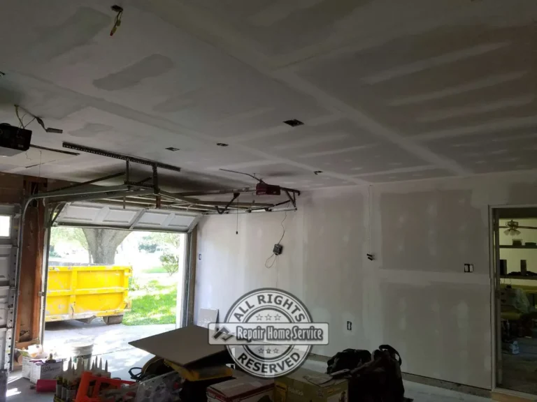 Garage drywall installation with taped ceiling seams, patched walls, open garage door, electrical boxes, tools, and debris container outside.