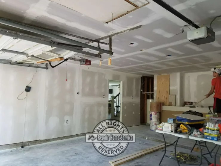 Garage Sheetrock installation in Houston TX with taped ceiling seams, patched walls, open garage door track, ceiling opener, and drywall panels ready for final finishing.